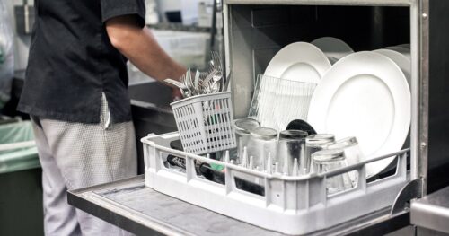 A man standing next to a opened dishwasher in a restaurant kitchen.